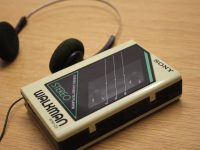 Ilustrasi pemutar kaset jinjing lawas beserta headphone di atas permukaan kayu.