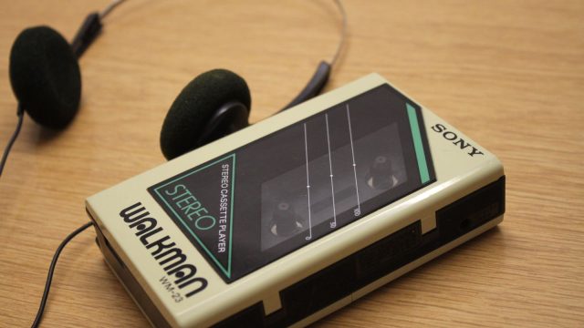 Ilustrasi pemutar kaset jinjing lawas beserta headphone di atas permukaan kayu.