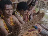 Perempuan adat Papua berkalung manik duduk berderet sambil merajut noken dari serat kayu