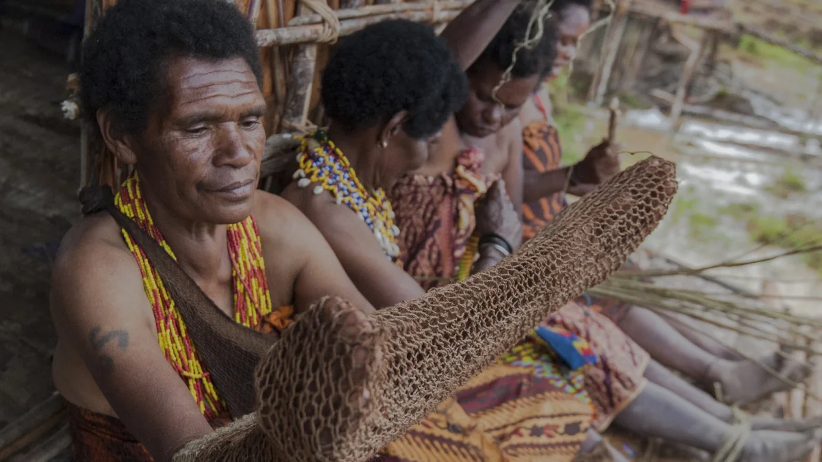 Perempuan adat Papua berkalung manik duduk berderet sambil merajut noken dari serat kayu