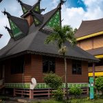 Ilustrasi rumah gadang beratap gonjong berdiri di antara pepohonan.