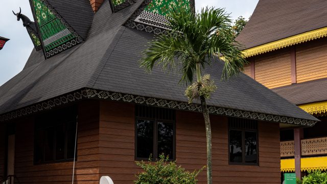 Ilustrasi rumah gadang beratap gonjong berdiri di antara pepohonan.