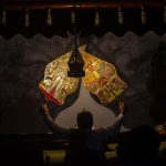 Dua wayang kulit berwarna keemasan berdiri di depan kelir yang disorot lampu dalam sebuah pertunjukan.
