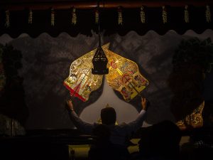 Dua wayang kulit berwarna keemasan berdiri di depan kelir yang disorot lampu dalam sebuah pertunjukan.