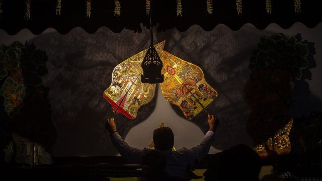 Dua wayang kulit berwarna keemasan berdiri di depan kelir yang disorot lampu dalam sebuah pertunjukan.
