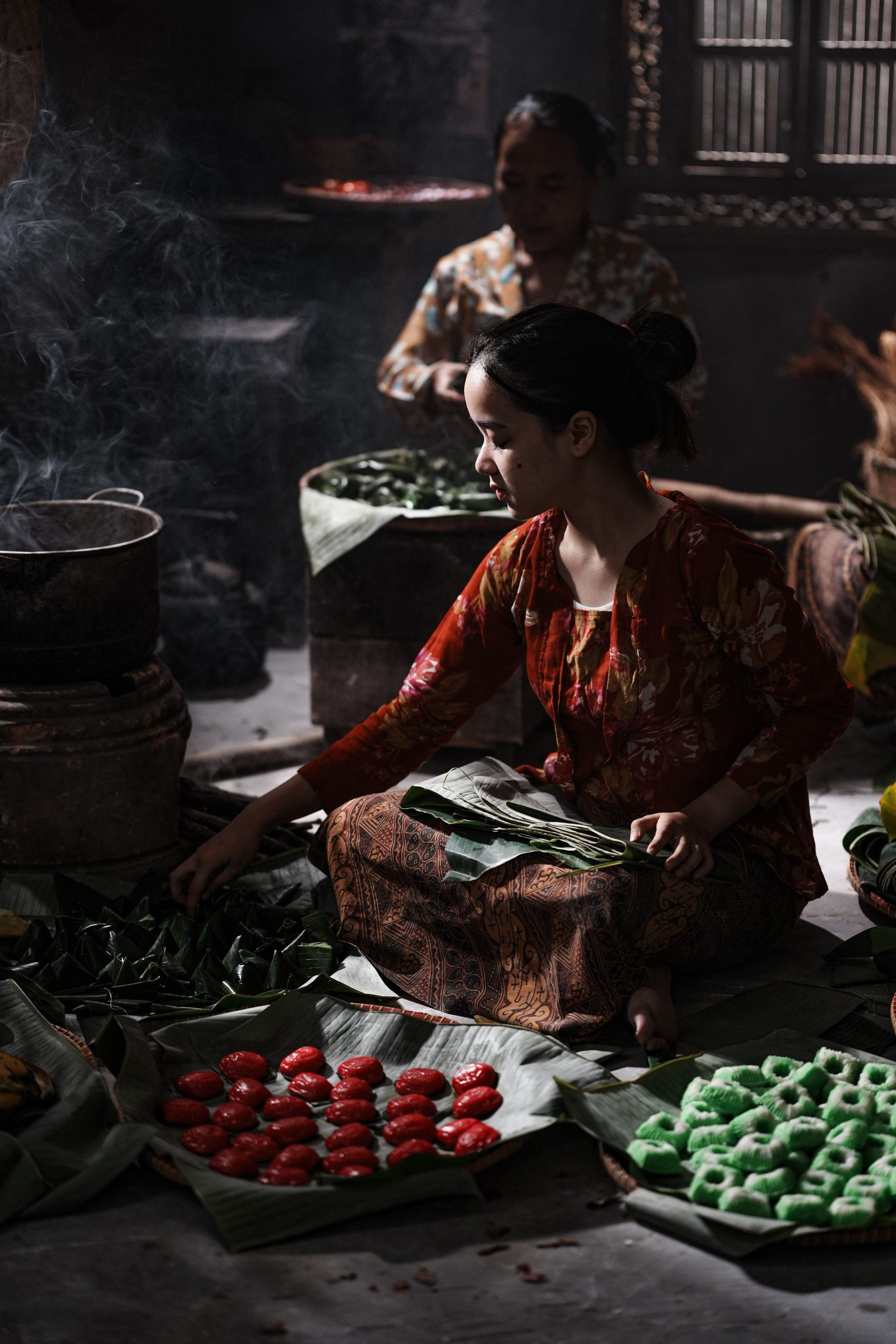 Ilustrasi seorang perempuan berkain batik menyiapkan makanan di dapur tradisional yang remang.