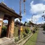 Ilustrasi jalan desa di Bali yang dihiasi penjor dengan gapura pura di kejauhan.