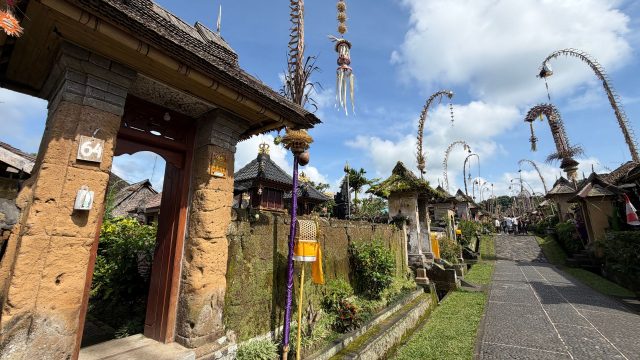 Ilustrasi jalan desa di Bali yang dihiasi penjor dengan gapura pura di kejauhan.