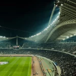 Tribun stadion dipenuhi penonton di bawah lampu terang pada malam hari