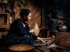 Perempuan menyalakan kayu bakar di tungku dapur tradisional berdinding anyaman bambu