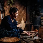 Perempuan menyalakan kayu bakar di tungku dapur tradisional berdinding anyaman bambu