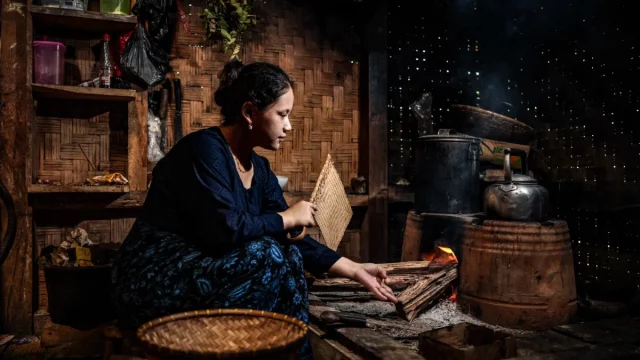 Perempuan menyalakan kayu bakar di tungku dapur tradisional berdinding anyaman bambu