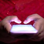Ilustrasi sepasang tangan mengetik di layar telepon genggam dalam ruangan gelap.