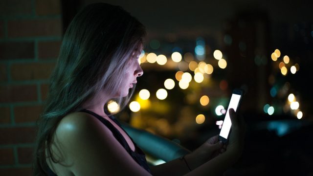 Ilustrasi seorang perempuan menatap layar telepon genggam pada malam hari.