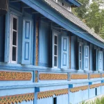 Dinding kayu masjid berwarna biru dengan panel ukiran bercat emas dan latar pepohonan