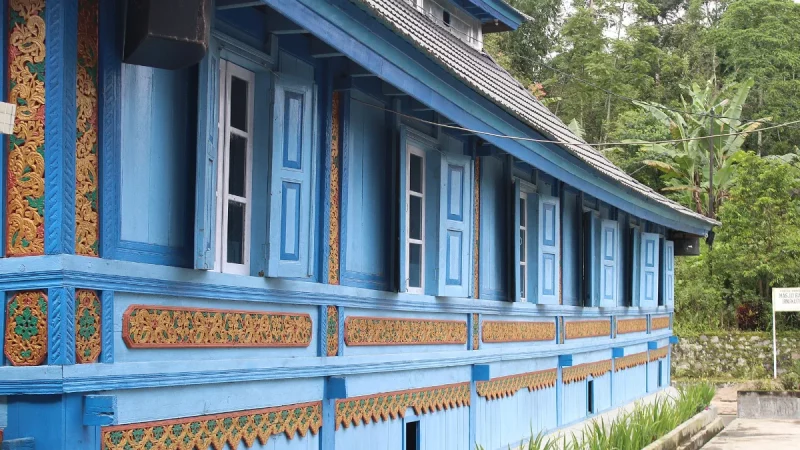 Dinding kayu masjid berwarna biru dengan panel ukiran bercat emas dan latar pepohonan