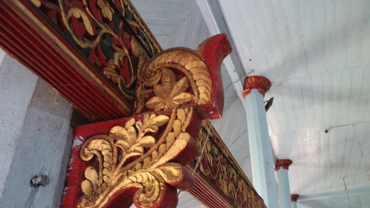 Detail ukiran kayu bermotif sulur bercat merah dan emas di bawah atap masjid