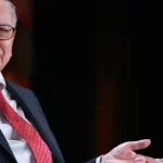 Warren Buffett mengenakan jas gelap dan dasi merah saat berbicara di sebuah acara