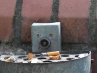 Ilustrasi asbak logam di dinding bata dengan sebatang rokok menyala yang mengepulkan asap