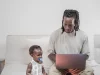 Ilustrasi: seorang ayah mengetik di laptop di sofa dengan anak balita di sampingnya