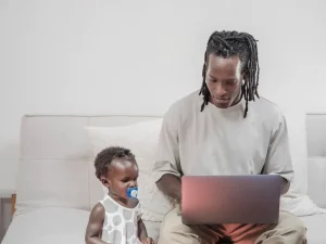 Ilustrasi: seorang ayah mengetik di laptop di sofa dengan anak balita di sampingnya