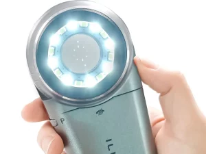 Ilustrasi tangan menggenggam dermatoskop dengan lingkar lampu LED menyala di sekeliling lensa