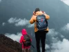 Ilustrasi dua pendaki beransel memandang gunung berselimut awan di Indonesia, tampak dari belakang