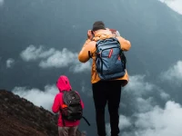 Ilustrasi dua pendaki beransel memandang gunung berselimut awan di Indonesia, tampak dari belakang