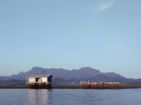 Ilustrasi rumah panggung dan keramba di atas laut dengan siluet Gunung Sepincan di Pulau Lingga, Kepulauan Riau