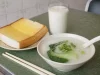 Ilustrasi semangkuk bubur sawi hijau, roti bakar mentega, dan segelas susu di meja kedai