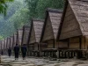 Ilustrasi deretan rumah kayu beratap rumbia dan warga berbaju hitam berjalan membelakangi