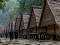 Ilustrasi deretan rumah kayu beratap rumbia dan warga berbaju hitam berjalan membelakangi