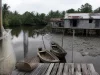 Ilustrasi dua sampan kayu tertambat di dermaga papan dekat rumah panggung beratap seng