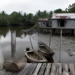 Ilustrasi dua sampan kayu tertambat di dermaga papan dekat rumah panggung beratap seng