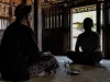 Ilustrasi lelaki tua berikat kepala duduk berhadapan dengan tamu di rumah kayu, segelas air dan sepiring garam di tikar
