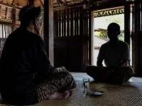 Ilustrasi lelaki tua berikat kepala duduk berhadapan dengan tamu di rumah kayu, segelas air dan sepiring garam di tikar