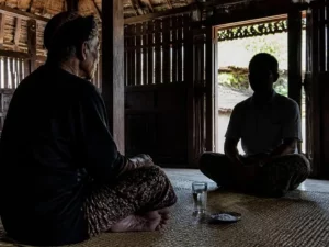 Ilustrasi lelaki tua berikat kepala duduk berhadapan dengan tamu di rumah kayu, segelas air dan sepiring garam di tikar