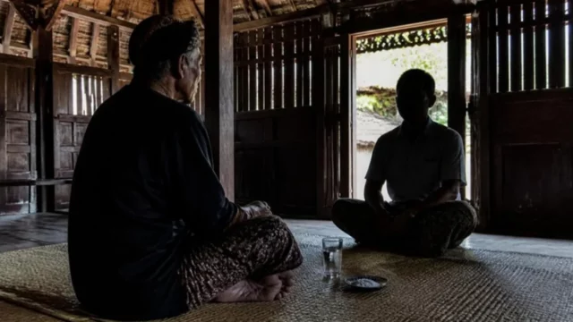 Ilustrasi lelaki tua berikat kepala duduk berhadapan dengan tamu di rumah kayu, segelas air dan sepiring garam di tikar