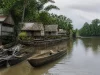 Ilustrasi perahu lesung kayu tertambat di muara berbakau dekat rumah panggung kampung pesisir