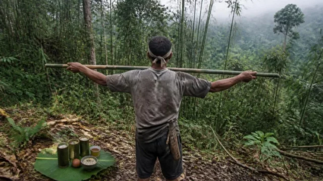 Ilustrasi peladang merentangkan sebatang bambu di tepi hutan berkabut, sesajen lemang dan telur di daun pisang