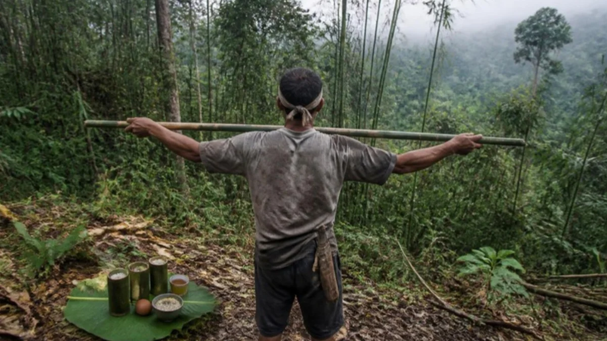 Ilustrasi peladang merentangkan sebatang bambu di tepi hutan berkabut, sesajen lemang dan telur di daun pisang
