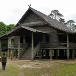 Ilustrasi rumah adat panggung kayu berserambi dan bertangga dengan dua orang berjalan di halaman