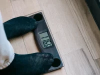 Ilustrasi sepasang kaki berdiri di atas timbangan badan digital di lantai kayu