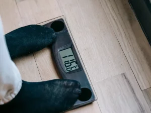Ilustrasi sepasang kaki berdiri di atas timbangan badan digital di lantai kayu