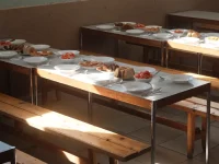 Ilustrasi kantin kosong dengan meja panjang berisi piring makanan dan bangku kayu tersinari matahari
