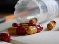 Ilustrasi kapsul antibiotik amoksisilin merah-kuning tumpah dari botol obat di atas meja