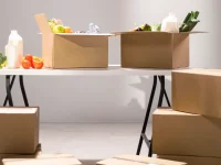 Ilustrasi kardus berisi sayur, buah, dan botol susu di atas meja lipat putih