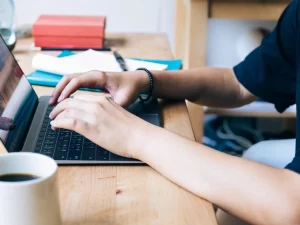 Ilustrasi perempuan muda mengetik di laptop di meja kayu dengan cangkir kopi dan buku catatan