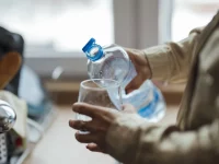 Ilustrasi tangan seseorang menuang air dari botol plastik ke gelas bening di dapur