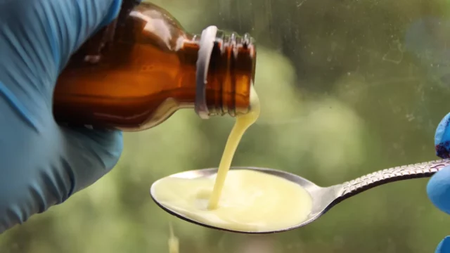 Ilustrasi tangan bersarung karet biru menuang obat cair dari botol ke sendok